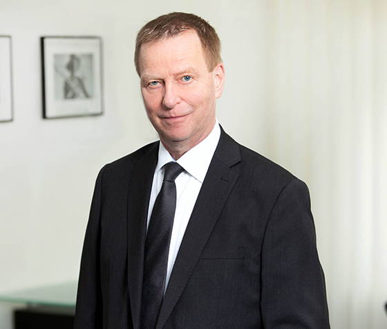 Mann im schwarzen Anzug und Krawatte steht drinnen vor weißer Wand mit gerahmten Bildern.