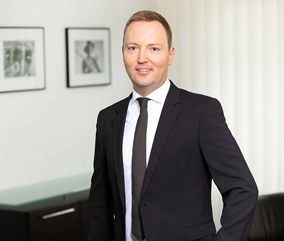 Mann im schwarzen Anzug und Krawatte steht im Büro vor gerahmten Fotos und weißen vertikalen Jalousien.