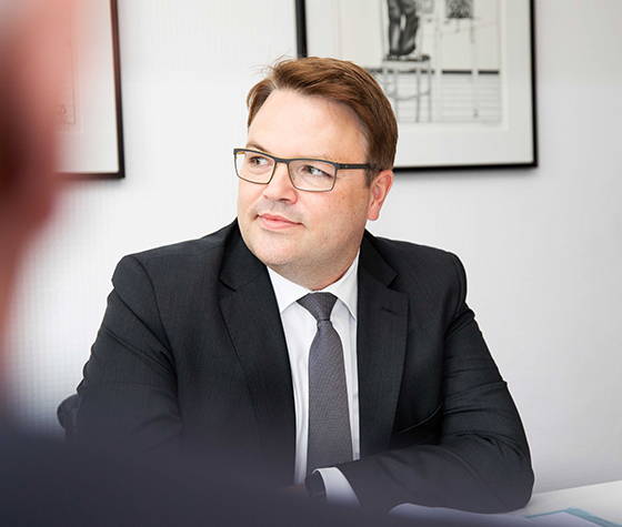 Mann im Anzug und mit Brille sitzt am Tisch und blickt zur Seite, hinter ihm hängen gerahmte Bilder an der Wand.